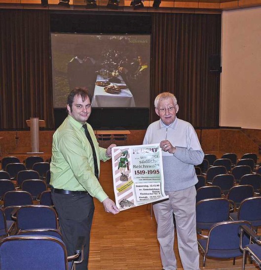 Willi Memmel und Alexander Hommel, 1. Vorsitzender des Arbeitskreises Feuchter Gewerbe und Hauptorganisator des Zeidlermarktes, zeigen das Plakat von der Erstaufführung des Films. Am 13. Mai wird „Feucht . . . und der südliche Reichswald 1519 – 1995“ in der Reichswaldhalle gezeigt. Im Hintergrund läuft Memmels Lebenswerk bereits zur Probe. Foto: Rüsing