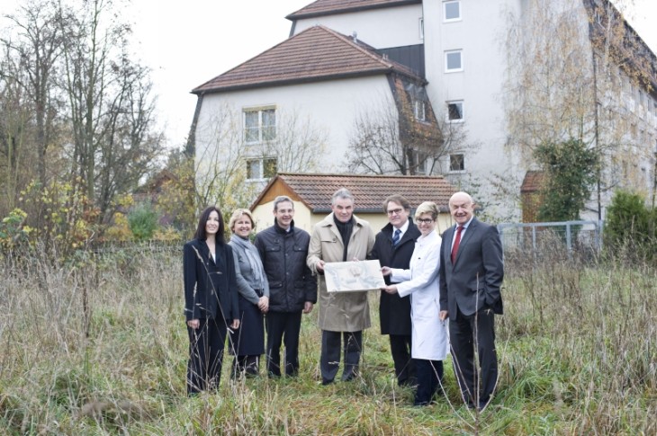 Auf dem Acker, auf dem diese sieben Menschen stehen, wird der Anbau entstehen. Im Hintergrund die alte PsoriSol-Klinik, die mit einem Übergang zum Neubau verbunden wird. Das Foto zeigt links: Carmen Adam,Prokuristin Marlene Greil, Bürgermeister Robert Ilg, Michael Hecke von der Besitzer-Gesellschaft, Architekt Professor Peter Haid, Chefärztin Clarissa Allmacher und Geschäftsführer Herbert Pichler.  Die Turnhallen-Insel wird von der Pegnitz umrahmt, Hochwasserschutzmaßnahmen waren deshalb nötig.