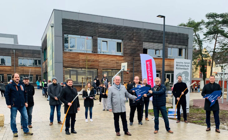 Die Geschwister-Scholl-Mittelschule (im Hintergrund) ist eine der Einrichtungen in Röthenbach, die an das Glasfasernetz angeschlossen wurden. Über die Fertigstellung freuen sich – unter anderem – Schulvertreter und Stadträte sowie Bürgermeister Klaus Hacker, Landrat Armin Kroder und der Laufer IT-Unternehmer Johannes Bisping.