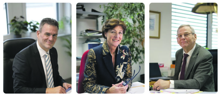 Auf dem Foto sieht man drei Personen, eine Frau und zwei Männer an ihren Schreibtischen