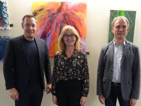 Auf dem Foto sieht man drei Personen; eine Frau und zwei Männer vor einer Bilderwand