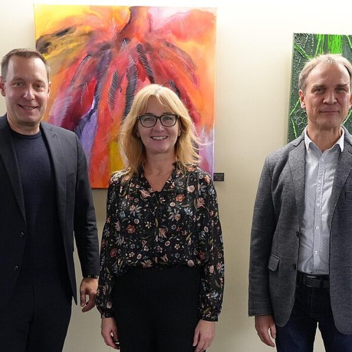 Auf dem Foto sieht man drei Personen; eine Frau und zwei Männer vor einer Bilderwand