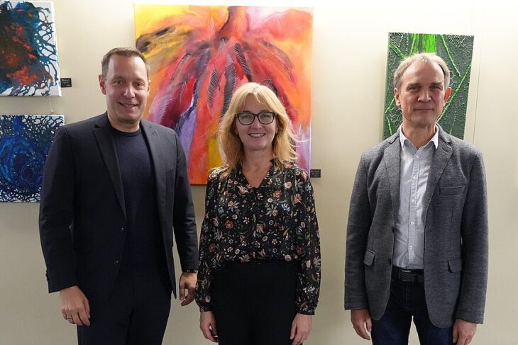 Auf dem Foto sieht man drei Personen; eine Frau und zwei Männer vor einer Bilderwand