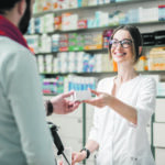 Eine junge Apothekerin händigt einem männlichen Kunden, der am Kassenschalter der Apotheke steht, Medikamente aus.