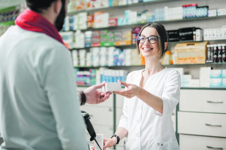 Eine junge Apothekerin händigt einem männlichen Kunden, der am Kassenschalter der Apotheke steht, Medikamente aus.