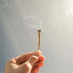 man-holding-a-lit-joint-of-marijuana-with-a-soft-sky-and-shiny-sun-in-the-background-cigarette-with-smoke-right-and-top-copy-space