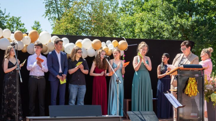 Schulleiterin Birgit Seidl (am Pult) zeichnet die besten Schülerinnen und Schüler des Jahrgangs aus (von links): Eleanor Gheorge, Rahoz Nuri, Moritz Krauß, Alexandru Mágurean, Thea Völkel, Antonia Paul , Milena Lukinskiy und Kristal Singh.