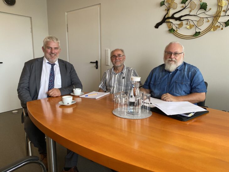 Auf dem Foto sieht man drei Personen an einem Tisch sitzen