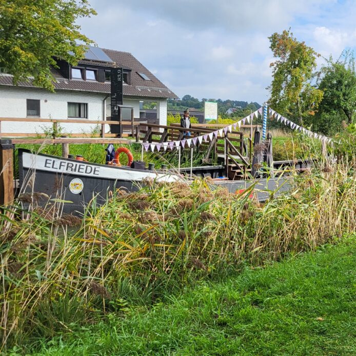 Das Treidelschiff Elfriede liegt am Gasthaus „Zum Ludwigskanal“ in Schwarzenbach vor Anker. Das Kaltblutpferd Fritz vom Landgasthof und Kutschbetrieb „Zum Ross“ in Hausheim grast gemütlich am Ufer, an dem viele Apfelbäume stehen.